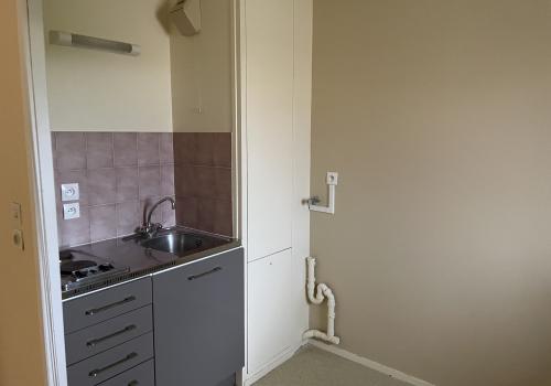 Coin cuisine avec évier et plaque de cuisson avec meuble de rangements gris en dessous avec une porte et 4 tiroirs. , Carrelage mural rose avec 2 prises au dessus de l'évier et lumière mural, à coté meuble beige de rangements, sol beige et murs beige et rosé.
