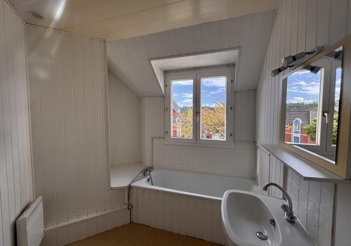 Salle de bain avec murs en lambris bois blanc, baignoire et lavabo avec son miroir, fenêtre à 2 battants au dessus de la baignoire.