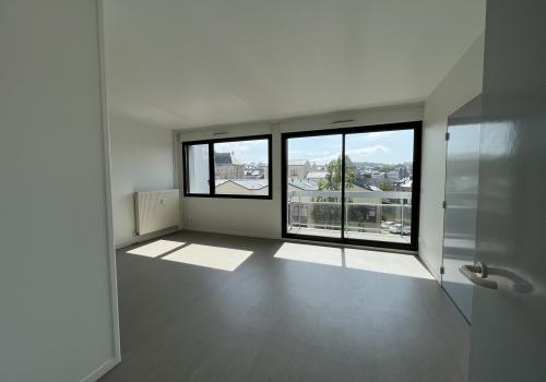 Pièce de vie avec une fenêtre simple ainsi qu'une baie vitrée donnant sur un balcon exposée plein sud. Le sol est en imitation parquet marron clair et le plafond et les murs sont blancs, une porte fermée grise est visible sur la droite. un radiateur mural est disposé sur le mur à gauche de la fenêtre.