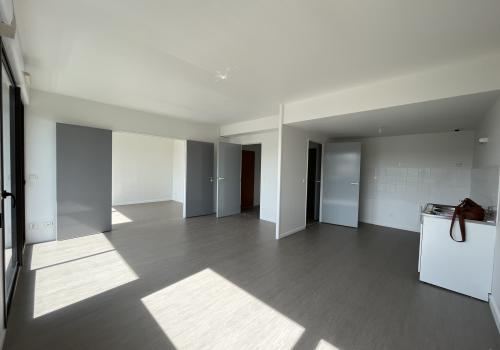 Vue sur l'ensemble de l'appartement, sol marrons clair ainsi qu'un espace cuisine est visible, de nombreuses portes grises claires donnant sur d'autres pièces sont également visibles et une porte-fenêtre apporte une belle luminosité à la pièce.